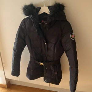 Svart pufferjacka med fuskpäls från Jackan.com - Svart pufferjacka från Jackan.com med avtagbar huva och fuskpäls. Jackan har bälte i midjan, dragkedja och knappar framtill samt emblem på ärmen. Perfekt för kalla dagar med sin quiltade design och långa ärmar.