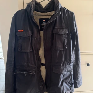 Svart parkajacka från Superdry - Stilren Field jacket från Superdry. Jackan har både dragkedja och knappar framtill, samt hög krage och emblem på ärmen. Perfekt för kalla dagar.