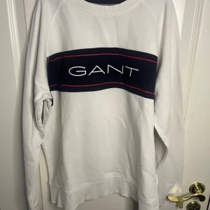 Vit sweatshirt från GANT med logga - Vit sweatshirt från GANT med en bred marinblå panel över bröstet och vit broderad logga. Röd detaljlinje längs panelen. Klassisk rund halsringning och långa ärmar. Mjuk bomullskänsla och relaxed fit, perfekt för en clean och sportig look. Storlek: 170 cm