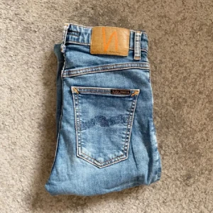 Blå jeans från Nudie Jeans, W26 L32 - Snygga blå jeans från Nudie Jeans med klassisk femficksdesign. Jeansen har en ljus tvätt och subtila slitningar, samt Nudie-logga på bakfickan och läderpatch i midjan. Perfekta för en avslappnad och trendig look. Hör av er vid frågor och funderingar!