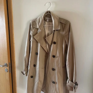 Beige trenchcoat med bälte - Klassisk beige trenchcoat med dubbelknäppning, bred krage och bälte i midjan samt vid ärmsluten. Jackan har slät yta och är helfodrad, perfekt för lager-på-lager-stil. Snygg och tidlös modell som funkar till allt.