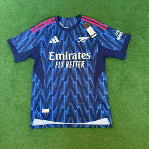 Arsenal 25/26 blå Adidas fotbollströja - Snygg Arsenal fotbollströja från Adidas i mörkblått med elektriskt blått blixtmönster. Tröjan har korta ärmar, röda Adidas-ränder på axlarna och klubbens logga på bröstet. Tillverkad i lätt och ventilerande polyester, perfekt för match eller träning.
