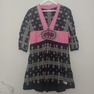 Odd Molly mönstrad kortklänning - Snygg kortklänning från Odd Molly med svart och rosa bas, vita broderade mönster och en broderad detalj framtill. Djup v-ringning och trekvartsärm. Perfekt för dig som gillar unika och färgstarka plagg med boho-vibe.