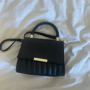 Svart handväska med gulddetaljer - Snygg svart handväska med quiltad nederdel och guldfärgade detaljer vid handtaget och låset. Väskan har ett stilrent och kompakt format med både handtag och axelrem.