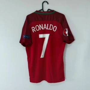 Portugal Ronaldo 7 EM 2016 Nike tröja  - Portugal fotbollströja från EM 2016 med Ronaldo 7 på ryggen. Röd med gröna och vinröda detaljer, officiella turneringsmärken på ärmarna och broderat landslagsmärke. Tillverkad i lätt och ventilerande Dri-FIT material. Perfekt för dig som älskar fotboll och samlartröjor.