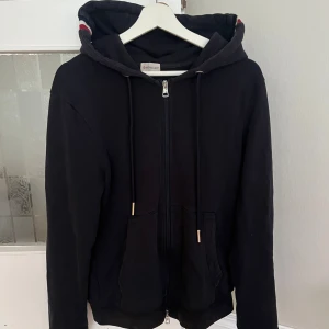 Marinblå hoodie från Moncler med dragkedja - Marinblå hoodie från Moncler med hel dragkedja, huva med röd och vit rand, samt logga på ärmen. Tröjan har långa ärmar, fickor framtill och ribbade muddar med röd och vit detalj. Perfekt för en chill och stilren look. Storlek L men sitter som M.  Pris kan diskuteras. 