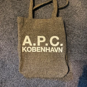 Brun totebag  från A.P.C. Köpenhamn - Snygg brun axelväska från A.P.C. Köpenhamn med fiskbensmönster och tydlig logga på framsidan. Väskan har långa axelband och är tillverkad i ett mjukt material som ger en chill och stilren vibe. Perfekt storlek för att bära med sig det viktigaste.