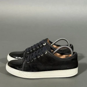 Svarta sneakers från Lanvin i mocka - Snygga svarta sneakers från Lanvin med ovandel i mocka och lackad tå. Storlek 40 men passar 41/42. Skorna har vita gummisulor och klassisk snörning. Perfekta för dig som gillar stilrena och exklusiva sneakers med en twist. Smutsiga och väldigt slitna på insidan. Inget og tillkommer 
