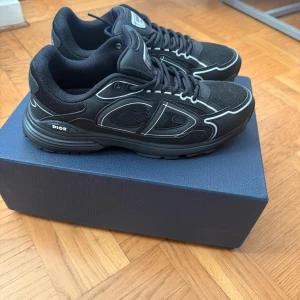 Svarta Dior B30 sneakers - Säljer ett par svarta Dior B30 sneakers med coola reflekterande detaljer och CD-logga på sidan. Skorna har mesh och syntetmaterial, snörning och en chunky sula. Perfekta för dig som vill sticka ut med en lyxig och sportig vibe.