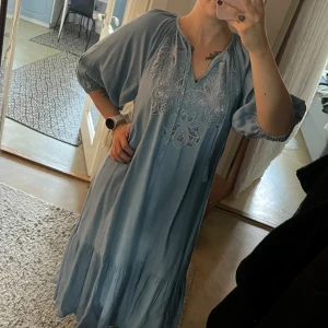 Blå klänning med broderi - Ljusblå långklänning med v-ringning och broderade blommor framtill. Klänningen har trekvartsärmar med resår och volang nertill. Tillverkad i mjukt Tencel-material som känns riktigt skönt mot huden. Prislapp kvar. Liten defekt men inget utmärkande  (visas i bild) 