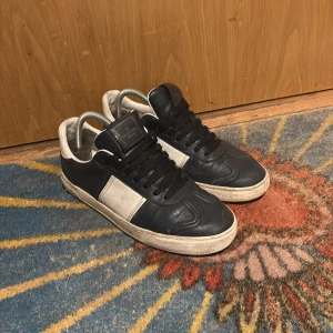 Valentino Garavani sneakers i svart och vitt - Snygga Valentino Garavani sneakers i svart och vitt skinn med klassisk låg profil och svarta skosnören. Skorna har en vit panel på sidan och diskret Valentino-logga på plösen. Perfekta för dig som gillar stilrena och exklusiva sneakers.