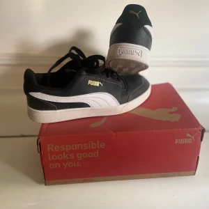 Svarta och vita sneakers från Puma - Snygga svarta sneakers från Puma med vita detaljer och guldlogga på sidan. Skorna har klassisk låg profil, snörning och vit sula. Ovandelen är i syntet och textil, perfekt för en sportig och stilren look. Bra skick inga skador