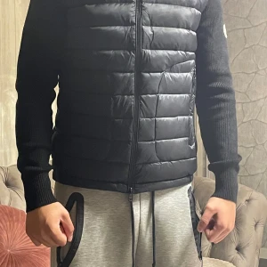 Svart Moncler cardigan med stickade ärmar - Säljer en svart cardigan från Moncler med quiltad front och stickade ärmar. Jackan har dragkedja framtill, två sidofickor med dragkedja och Moncler-logga på vänster ärm. Perfekt för höst och vinter, riktigt snygg och stilren modell.