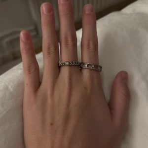 Två silvriga ringar med stenar - Säljer två snygga silvriga ringar. Den ena har ett kedjemönster med små infattade stenar, den andra har ett öppet rutmönster med glittriga detaljer runt om. Perfekta för att mixa och matcha till din stil.