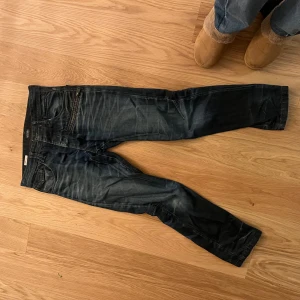 Mörkblå jeans från Jack & Jones - Snygga mörkblå jeans från Jack & Jones med slitna detaljer och dragkedja på fickan. Jeansen har knäppning med knappar och klassisk femficksdesign. Materialet är robust denim och passformen är rak med något smalare ben.