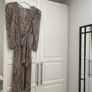 Zebramönstrad klänning från Adoore -  klänning från Adoore med zebramönster i beige och brunt. Klänningen har långa ärmar, markerad midja med knytband och lätt puff vid axlarna. Storlek 36