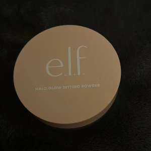 e.l.f. Halo Glow Setting Powder Light - Säljer ett e.l.f. Halo Glow Setting Powder i färgen Light. Det är ett löspuder med en ljus, neutral ton som ger en glowig finish. Kommer i en rund burk med beige lock och transparent underdel. Perfekt för att fixera sminket och ge huden en fräsch lyster.