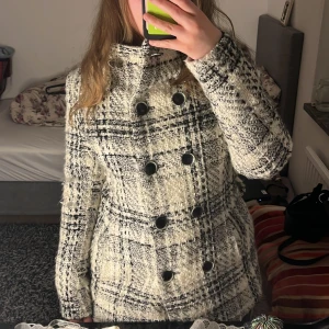 Rutig kappa från Zara Basic - Säljer en snygg rutig kappa från Zara Basic i svart och vitt. Jackan har dubbelknäppning med stora svarta knappar och hög krage. Materialet är grovt och känns ulligt, perfekt för kyligare dagar. 