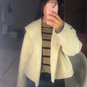 Krämvit/ beige kappa - Säljer en krämvit kappa med oversized passform och stora slag. Jackan har två svarta knappar framtill och snedställda fickor. Den är tillverkad i ett ull-liknande material och har lång ärm.