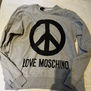 Grå sweatshirt från Love Moschino - Säljer en grå sweatshirt från Love Moschino med stort svart peace-märke och text framtill. Tröjan har rund halsringning och långa ärmar. Perfekt för dig som gillar statement-plagg och vill sticka ut lite extra.