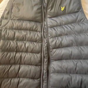 Svart dunväst från Lyle & Scott - Snygg svart dunväst från Lyle & Scott med klassisk gul logga på bröstet. Västen har horisontella quiltade partier och dragkedja framtill. Perfekt för lager-på-lager och enkel att matcha med olika outfits.