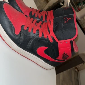 Nike Air Jordan 1 High svart/röd 45.5 - Säljer ett par Nike Air Jordan 1 High i klassisk svart och röd färg. Skorna har röd swoosh, röda snören och detaljer, samt svart läder på ovandelen. Ikonisk Jordan-logga på sidan och tungans etikett. Perfekta för dig som gillar streetwear och sneakerskultur.