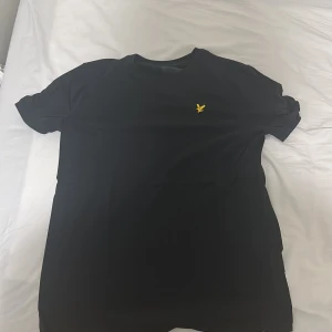 Svart t-shirt från Lyle & Scott - Svart t-shirt från Lyle & Scott med klassisk passform och korta ärmar. På bröstet sitter den ikoniska gula fågel-loggan som ger en clean och stilren look. Tillverkad i mjuk bomull som känns skön mot huden.