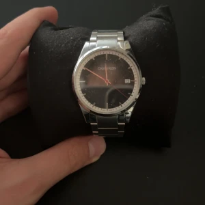 Silverfärgad Calvin Klein klocka - Stilren klocka från Calvin Klein med svart urtavla, röd sekundvisare och datumvisning. Behöver bara batterier Klockan har en rund form och ett klassiskt armband i silverfärgat metallmaterial. Perfekt för dig som gillar minimalistisk design och snygga detaljer.