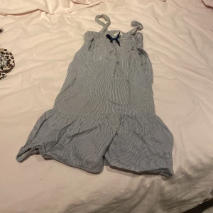 Randig klänning med rosett - Supersöt klänning med smala blå och vita ränder, volangkant nertill och en liten blå rosett framtill. Perfekta för varma dagar och har en luftig, loose passform. Axelbanden ger en unik look och materialet känns lätt och bekvämt.