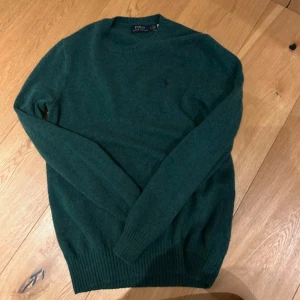 Mörkgrön stickad tröja Polo Ralph Lauren - Mörkgrön stickad tröja från Polo Ralph Lauren med klassisk rund hals och diskret broderad logga på bröstet. Tröjan har långa ärmar och ribbade muddar vid ärmslut och nederkant. Perfekt för lager-på-lager under höst och vinter. Flitigt använd och väldigt väldigt bra sick overall dessvärre finns det ett pyttelitet hål, går att fixa på cirka 10 minuter. 