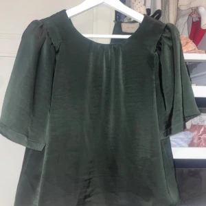 Mörkgrön blus från Neo Noir - Säljer en elegant mörkgrön blus från Neo Noir med rund halsringning och halvlånga, lätt puffade ärmar. Blusen har en lyxig glansig finish och är tillverkad i ett slätt material som ger en exklusiv känsla.