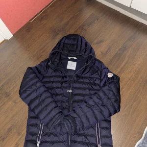 Mörkblå dunjacka från Moncler - Snygg mörkblå dunjacka från Moncler med huva och dragkedja framtill. Jackan har klassisk quiltad design, Moncler-logga på ärmen och två fickor med dragkedja. Perfekt för kyliga dagar och riktigt stilren look.