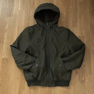 Snygg Olivgrön vindjacka  - Snygg Olivgrön vindjacka med huva och dragsko, hel dragkedja framtill och två fickor med tryckknappar. Jackan har ribbade muddar vid ärmslut och nederkant, perfekt för en stilren och avslappnad look.