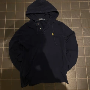 Mörkblå hoodie från Polo Ralph Lauren - Snygg mörkblå hoodie från Polo Ralph Lauren med klassisk gul logga på bröstet. Tröjan har huva med snörning, knappar vid halsen och är tillverkad i mjuk bomull. Perfekt för dig som gillar stilrena och bekväma plagg.