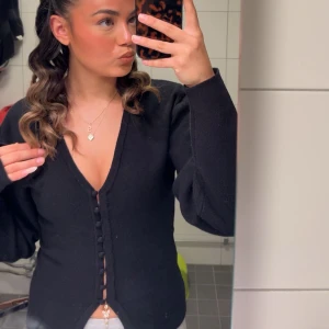 Svart kofta med v-ringning och knappar - Säljer en svart kofta med djup v-ringning och dekorativa knappar framtill. Koftan har långärmade puffärmar och är figurnära i modellen. Perfekt att styla med smycken för en trendig look.