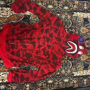 Röd camo hoodie från Bape - Säljer en ikonisk röd camo hoodie från Bape med hajmotiv på huvan och dragkedja hela vägen upp. Hoodien har långa ärmar och coola detaljer som broderade bokstäver och tänder på huvan. Perfekt statement-plagg för streetwear-stilen. Storlek XL men passar som L efter som man ska size upp