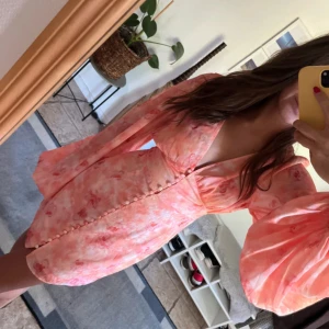 Adoore klänning - Superfin kortklänning i rosa och ljusa toner med blommigt mönster. Helt ny bara använt en gång! Djup v-ringning, markerad midja och knappar framtill. Klänningen har långa puffärmar som ger en romantisk vibe. Perfekt för sommarens alla tillfällen!