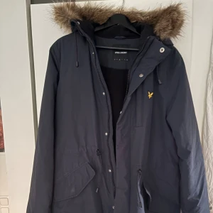 Mörkblå parkas från Lyle & Scott - Parkas från Lyle & Scott i mörkblå färg med huva och gul logga på bröstet. Jackan har dragkedja, knappar och snörning i midjan samt stora fickor framtill. I bra skick, perfekt för kyliga dagar. Har kemtvättats för inte så länge sen. Passar M/L, pälsen är avtagbar.