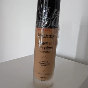 IsaDora Wake Up the Glow Foundation - Wake Up the Glow foundation från IsaDora i en beige nyans. Ger en naturligt glowig finish och innehåller hyaluronsyra och vitamin E. Kommer i en stilren glasflaska med svart pump. SPF 50 och vegansk formula. Nr 7N