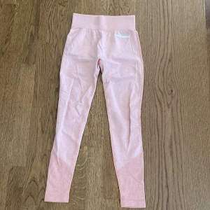 Rosa leggings från FUNGWEAR - Snygga ljusrosa leggings från FUNGWEAR med hög midja och ribbad linning. De har diskreta mönstrade detaljer längs benen och bak, samt logga framtill och baktill. Perfekta för träning eller chill, med stretchigt och mjukt material som sitter skönt. 