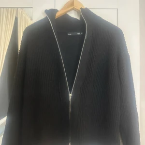 Svart stickad kofta med dragkedja - Svart stickad kofta från 1951 med ribbad struktur och hel dragkedja framtill. Koftan har långärmade och en enkel, stilren design som passar perfekt till lager-på-lager. Snygg att ha öppen eller stängd beroende på humör.