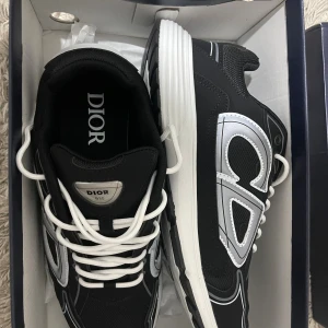 Svarta Dior B30 sneakers med vita detaljer - Säljer ett par svarta Dior B30 sneakers med vita och silverfärgade detaljer. Skorna har en chunky vit sula, tydlig Dior-logga på sidan och vita snören. Ovandelen är i mesh och syntetmaterial för en sportig vibe. Perfekta för dig som vill sticka ut med lyxiga sneakers. Skorna är aldrig använda. Kom gärna med pris förslag