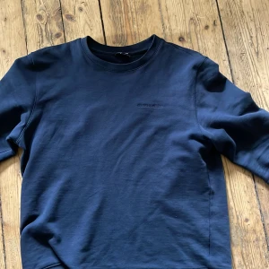 Mörkblå sweatshirt från J.Lindeberg - Säljer en stilren mörkblå sweatshirt från J.Lindeberg i bomull. Tröjan har rund halsringning, broderad logga på bröstet och långa ärmar. Perfekt för dig som gillar enkel och clean stil.
