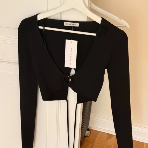 Svart croppad bolerotopp från Chiquelle  - Snygg svart croppad topp från Dilvin (Chiquelle)  med lång ärm och ribbad struktur. Toppen har en cool ringdetalj framtill och knytband som hänger ner. Perfekt för dig som gillar trendiga och stilrena plagg med lite edge. Står One Size men skulle säga att den passar S. 