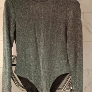 Glittrig silver body med öppen rygg - Säljer en glittrig silverfärgad body med lång ärm och djup v-ringning i ryggen. Materialet är stretchigt och skimrar snyggt i ljuset, perfekt för att sticka ut. Kroppsnära passform och hög hals framtill. Supersnygg till höga jeans eller kjol!