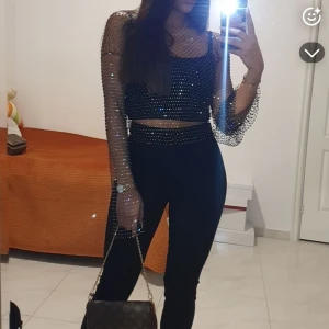 Svart glittrig croppad mesh-top - Säljer en svart croppad mesh-top med glittriga detaljer och långa transparenta ärmar. Toppen har en figurnära passform och är täckt av små glittrande stenar som ger en riktigt snygg effekt. Perfekt för att sticka ut med en cool och trendig look.