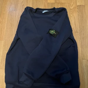 Mörkblå Stone Island sweatshirt - Säljer en mörkblå sweatshirt från Stone Island med den klassiska loggan på ärmen. Tröjan har rund hals, ribbade muddar och är tillverkad i mjuk bomull. Perfekt för dig som gillar streetwear och vill ha en clean look. Passar killar och tjejer pris kan diskuteras fraktar samma dag 