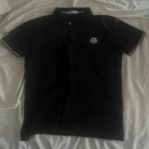 Svart Moncler pikétröja M - Svart pikétröja från Moncler i storlek M. Klassisk design med korta ärmar, krage och knappar framtill. Moncler-logga på bröstet och diskreta rödvita detaljer på ärmsluten. Perfekt för en clean och stilren look.