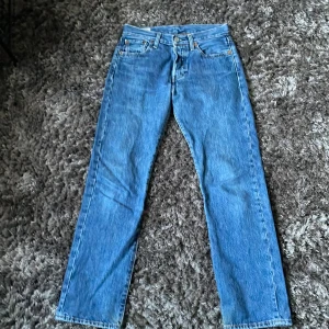 Levi's 501 blå jeans W27 L30 - Klassiska Levi's 501 jeans i blå denim med raka ben och normal passform. Byxorna har fem fickor, knappgylf och den ikoniska läderpatchen bak i midjan. Perfekta för dig som gillar tidlös stil och vill ha ett par äkta Levi's i garderoben.