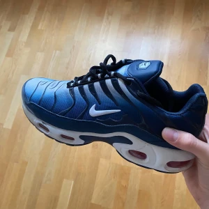 Nike Air Max Plus blå sneakers - Säljer ett par Nike Air Max Plus sneakers i blått med svarta och vita detaljer. Skorna har den klassiska Tn-loggan bak, vågiga linjer på ovandelen och en chunky vit sula med synliga luftbubblor. Ovandelen är i syntetmaterial och mesh för en sportig vibe. Perfekta för dig som gillar streetwear. Det står att skorna är storlek 42 inuti skorna, men det är egentligen storlek 41. Passar även för 40,5